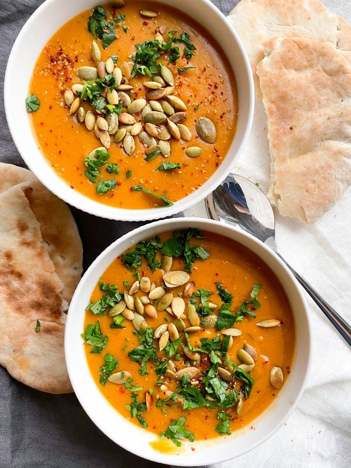 2 bowls of creamy lentil soup topped with toasted pumpkin seeds, chopped fresh herbs and red pepper flakes. Pitta bread on each side of the soup bowls