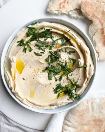 overhead image of smooth silky hummus in a flat bowl decorated with golden olive oil, chopped parsley and cracked black pepper. there is broken pieces of toasted pitta bread laying next to the hummus bowl on a linen towel