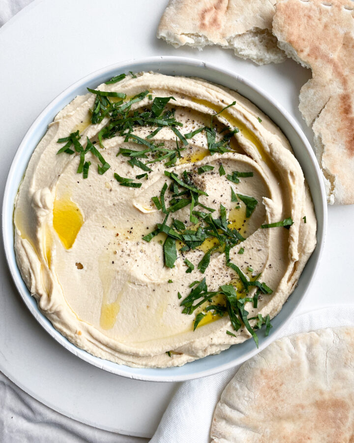 overhead image of smooth silky hummus in a flat bowl decorated with golden olive oil, chopped parsley and cracked black pepper. there is broken pieces of toasted pitta bread laying next to the hummus bowl on a linen towel